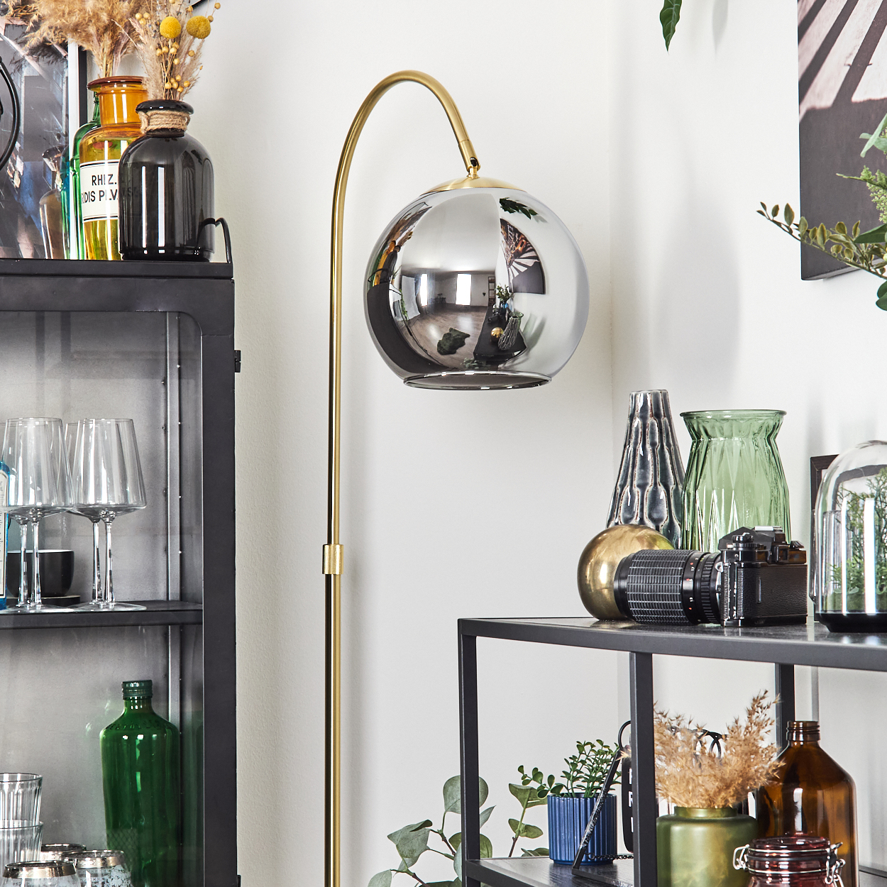 Eine goldene Lampe mit einem verspiegelten Kugelschirm ist der Blickfang. Regale zeigen Vasen, eine Kamera und andere Dekorationsartikel.