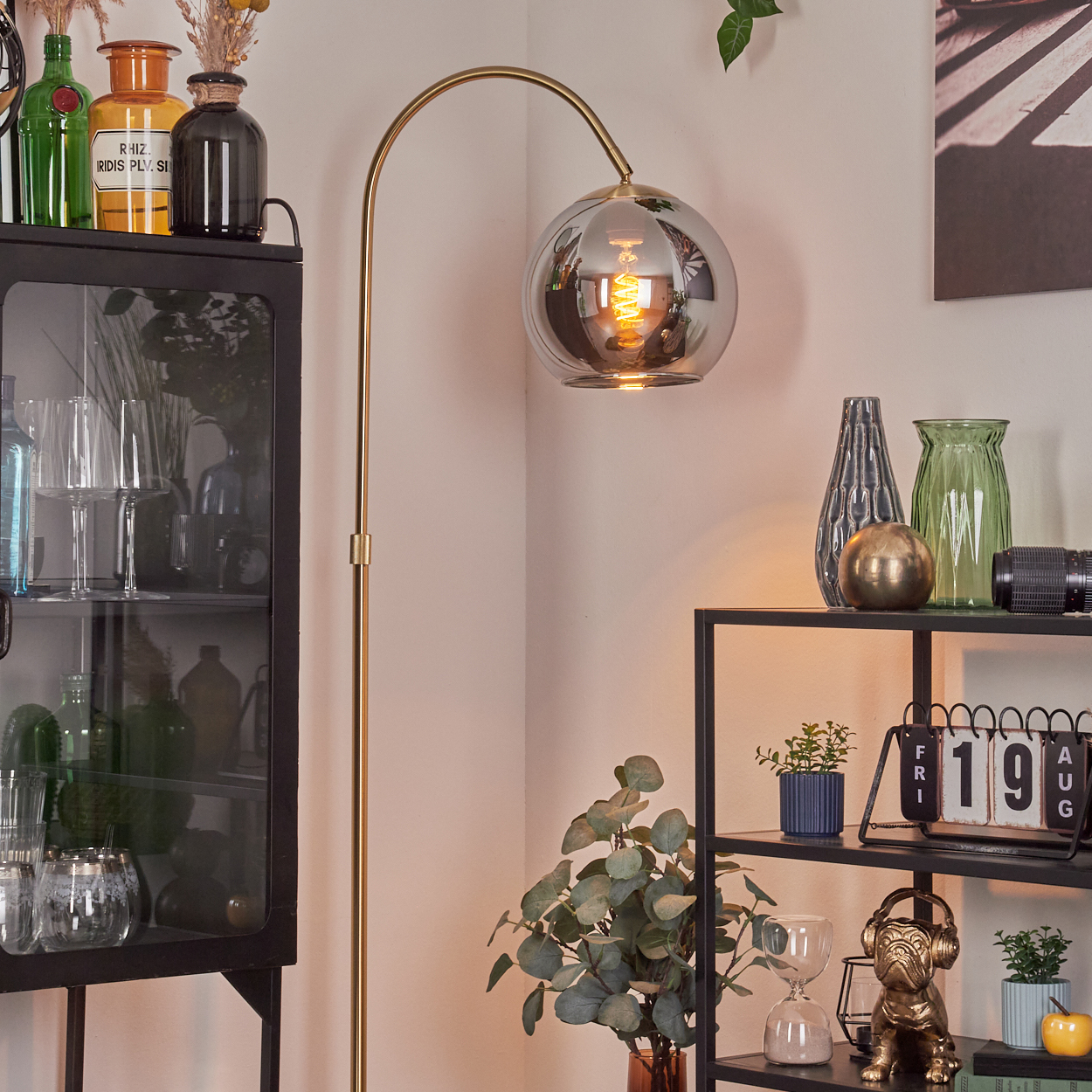 Eine goldene Stehlampe mit Glasschirm neben einem schwarzen Regal mit Vasen und Dekorationen.