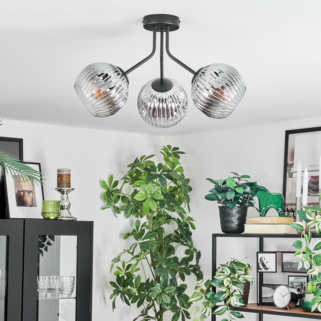 Eine moderne Deckenlampe mit drei Glasschirmen. Der Raum hat Pflanzen, schwarze Möbel und gerahmte Bilder.