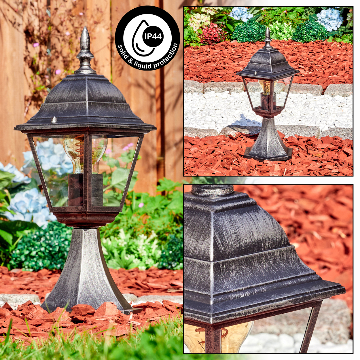 Eine dekorative Gartenlampe mit silbernem Sockel, Glasscheiben und einer Glühbirne. Sie steht in einem Gartenbeet mit roter Mulche und Pflanzen.