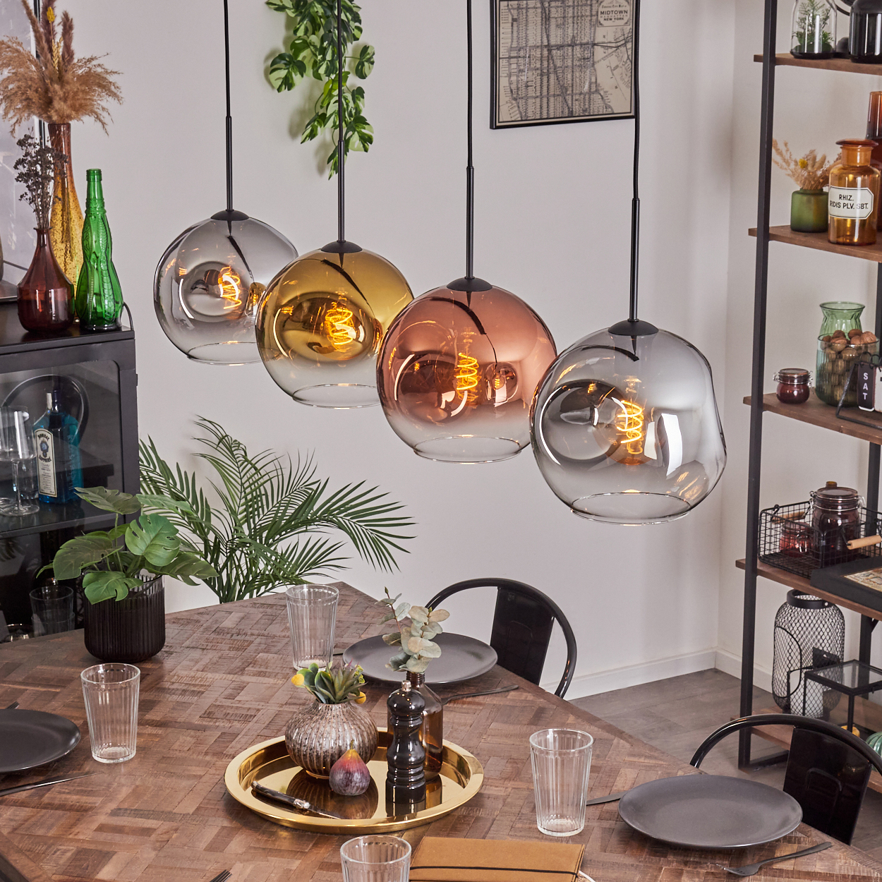 Vier Glas-Pendelleuchten hängen über einem Holztisch, der für das Abendessen gedeckt ist. Pflanzen, Regale mit Gläsern und eine Karte sind im Hintergrund.