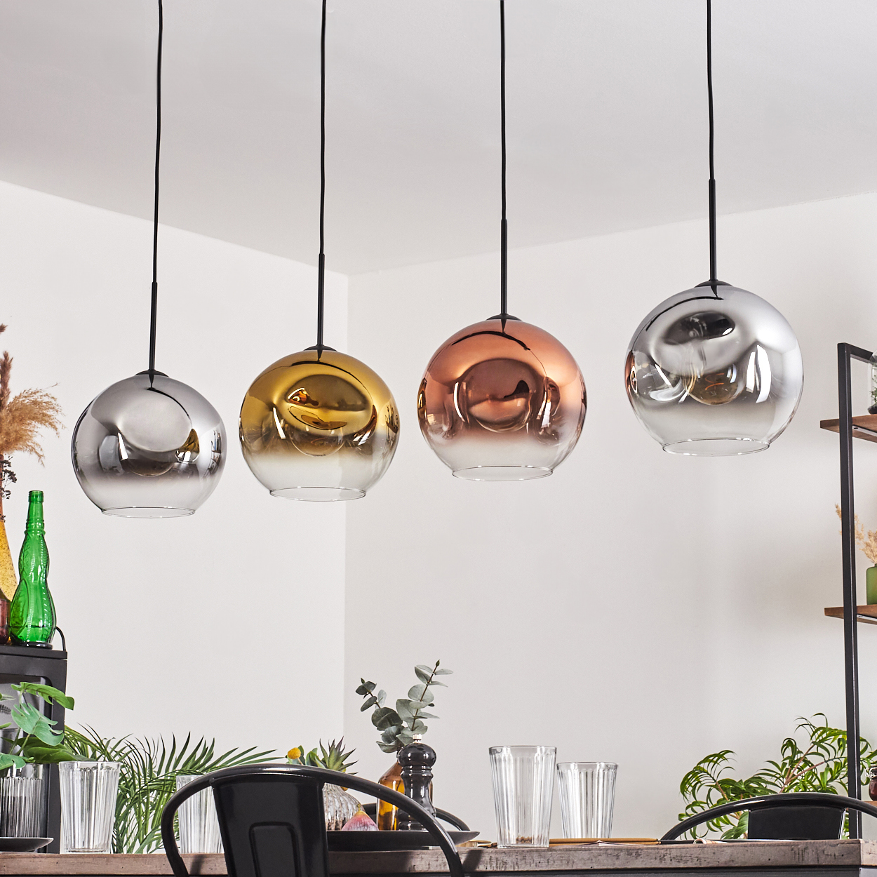 Vier kugelförmige Pendelleuchten in Silber, Gold, Kupfer und Silber hängen über einem Esstisch. Der Hintergrund ist weiß.
