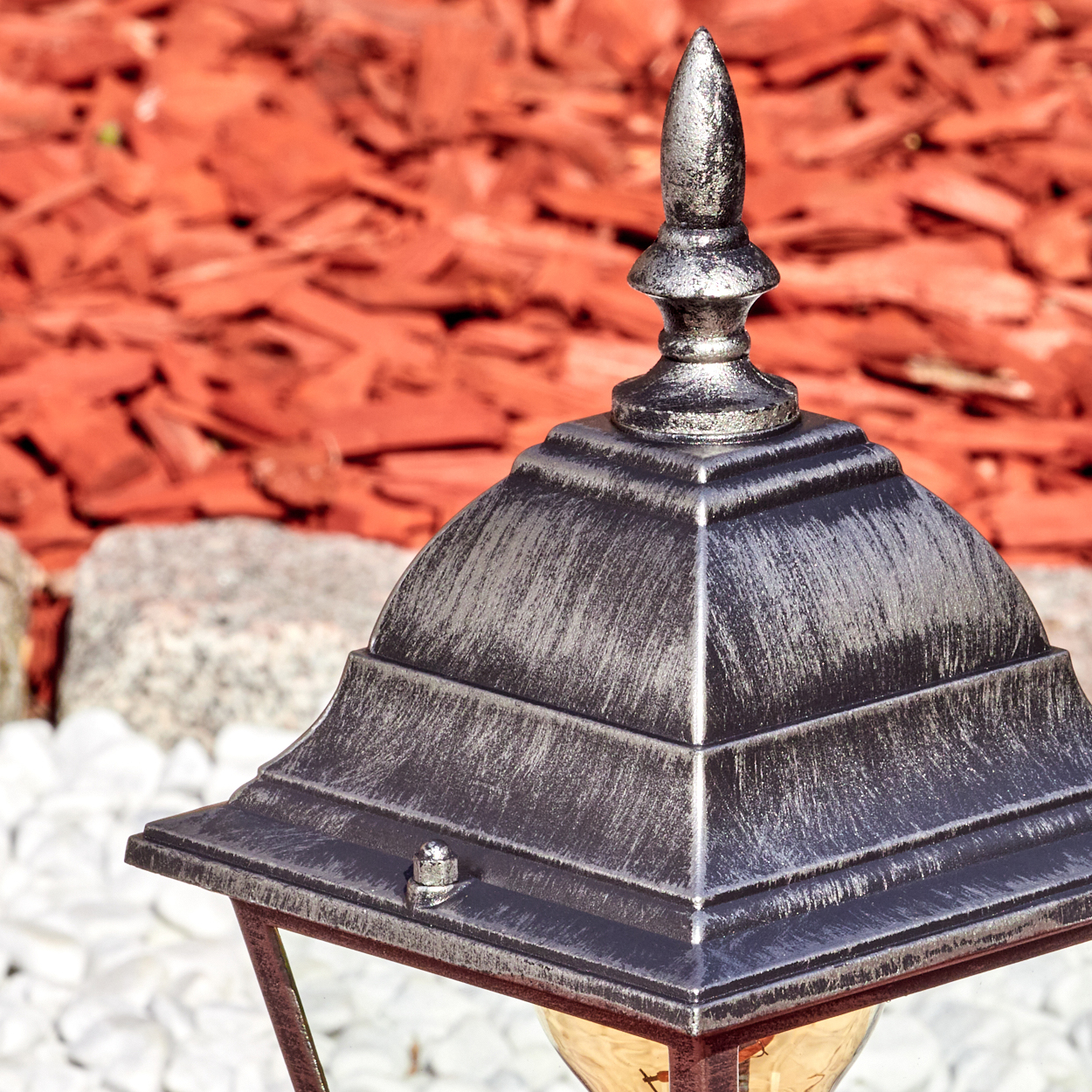 Eine dekorative Außenlampe mit silberner Spitze. Im Hintergrund roter Mulch und weiße Steine.