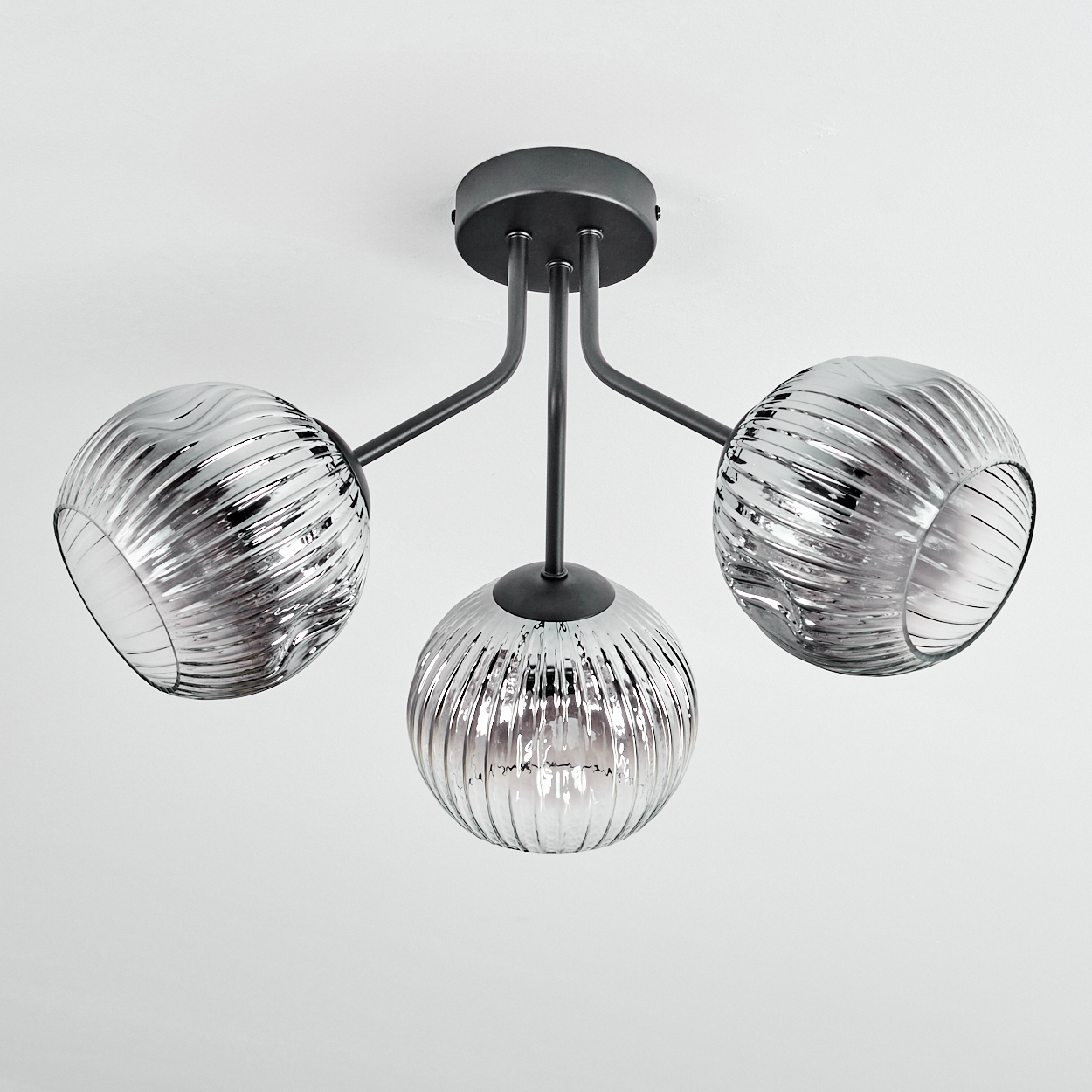 Eine moderne Deckenlampe mit drei kugelförmigen, geriffelten Glasschirmen. Die Halterung ist dunkelgrau und befindet sich vor weißem Hintergrund.