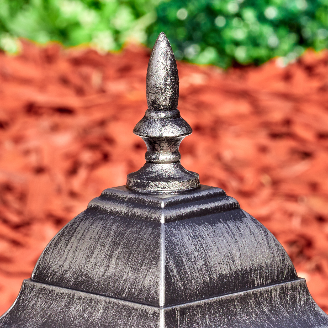 Nahaufnahme einer dekorativen Metallstruktur mit spitzer Spitze. Roter Mulch und grünes Laub im verschwommenen Hintergrund.
