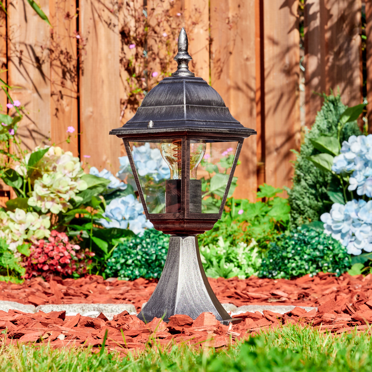 Eine Außenlampe in einem Garten mit Blumen und einem Holzzaun. Die Lampe ist schwarz und silber, und der Garten hat roten Mulch.