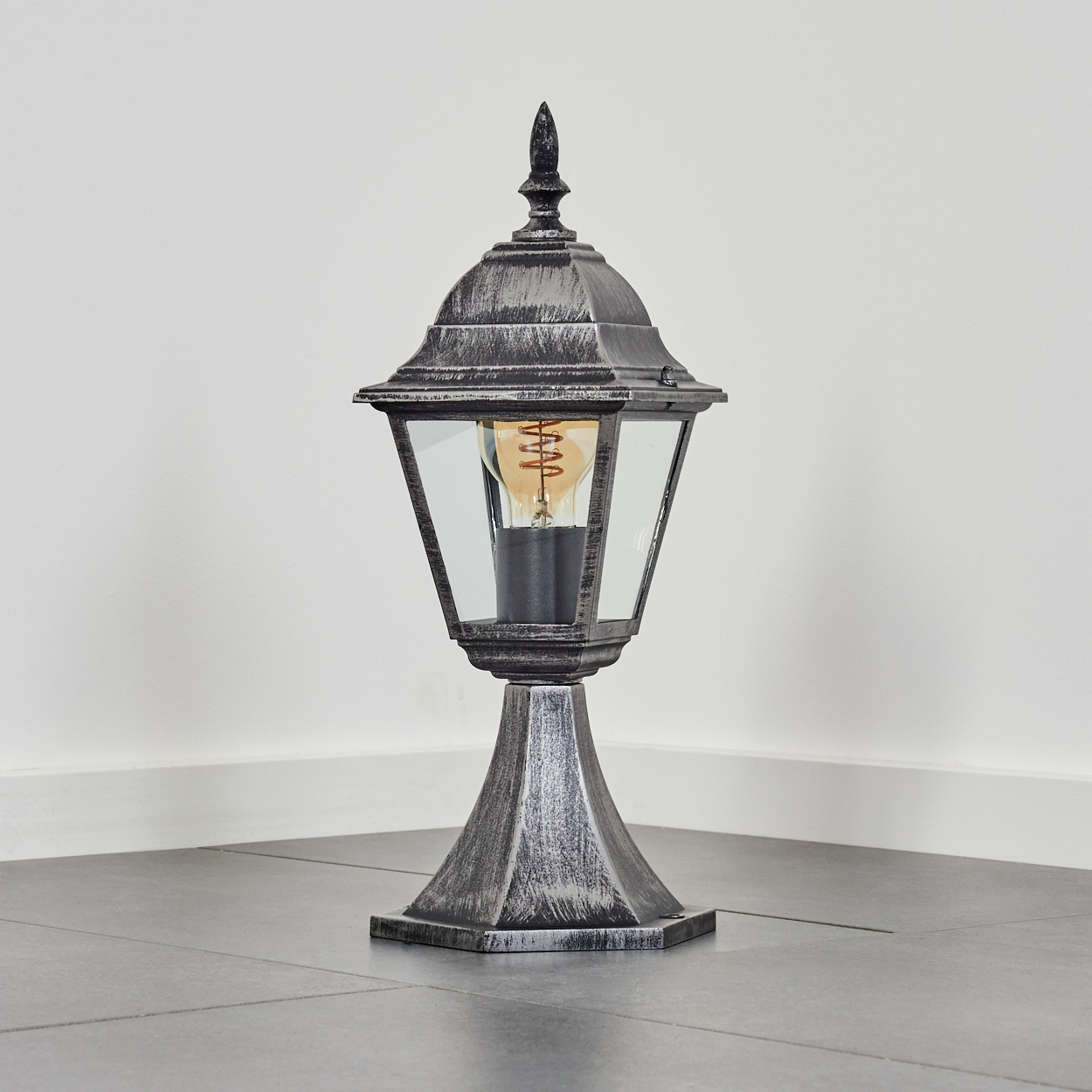 Eine Außenlampe steht auf einem gefliesten Boden. Sie hat einen dekorativen Metallrahmen mit Glasscheiben und eine leuchtende Glühbirne.