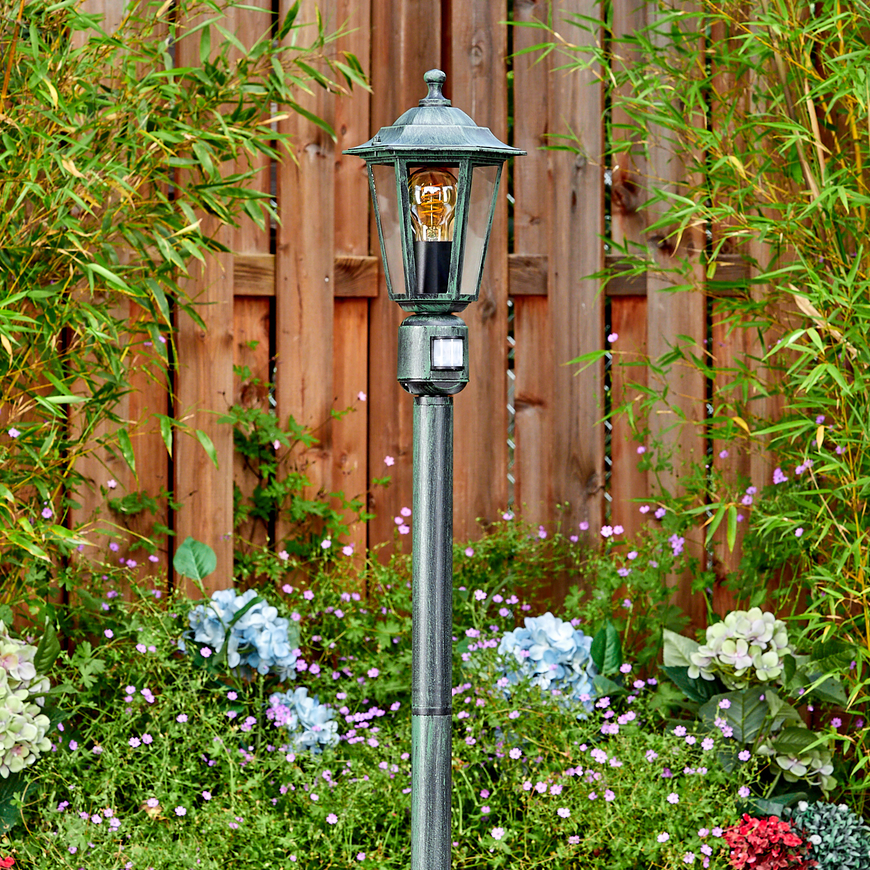 Eine dekorative Außenlampe mit einer Glühbirne steht in einem Garten mit Bambus und Blumen vor einem Holzzaun.