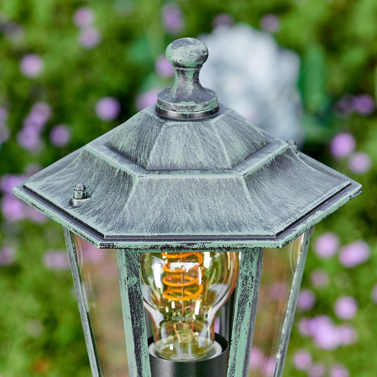 Nahaufnahme einer Vintage-Außenlampe mit grün-grauer Metalloberfläche und sichtbarer, leuchtender Glühbirne vor verschwommenem grünem Hintergrund.