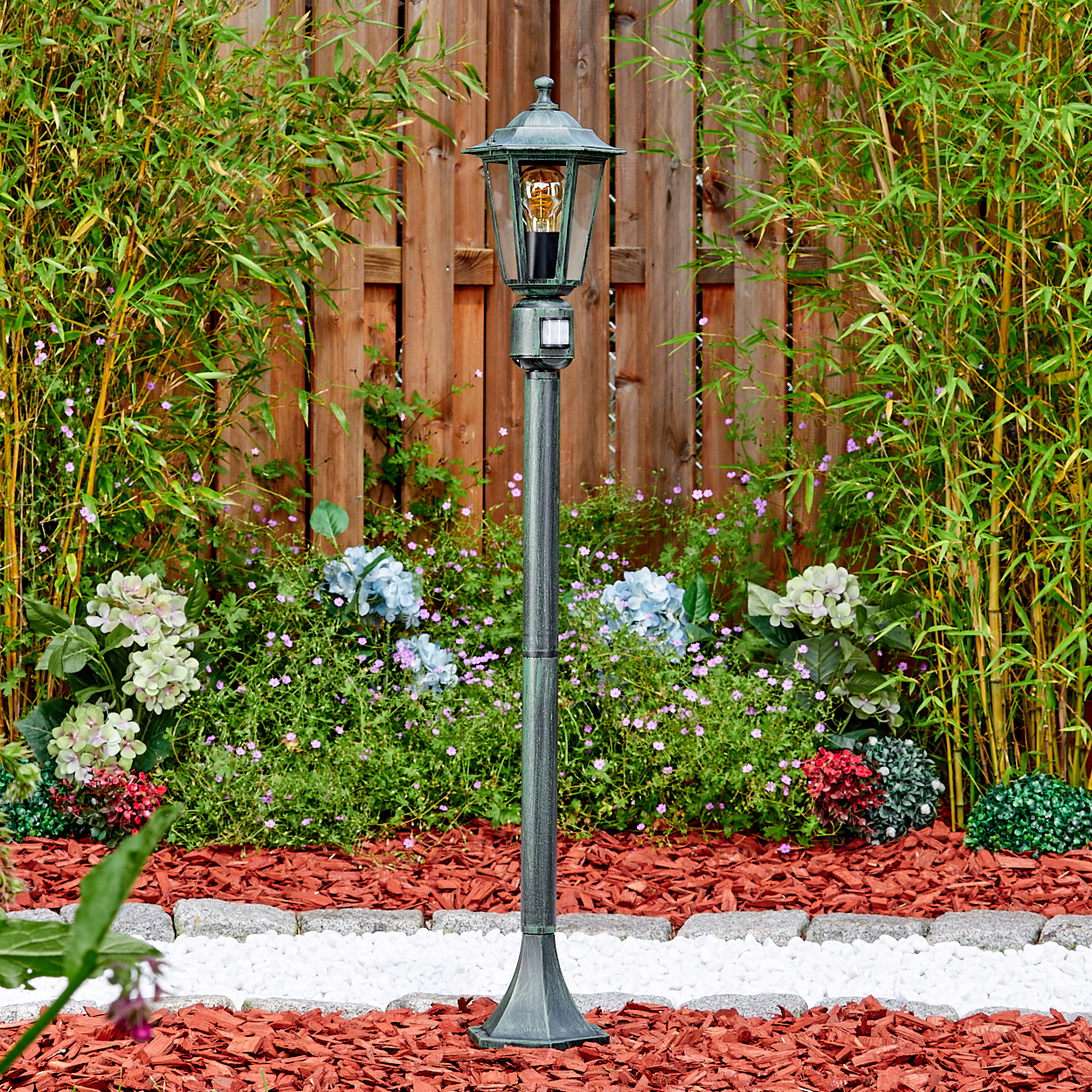 Eine dekorative Gartenlampe steht vor einem Holzzaun, umgeben von Pflanzen und Blumen. Rindenmulch bedeckt den Boden.