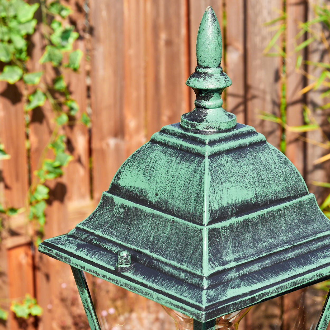 Nahaufnahme einer grünen, verzierten Außenlampe mit Holzzaun und Efeu im Hintergrund. Die Lampe ist detailliert und verwittert.