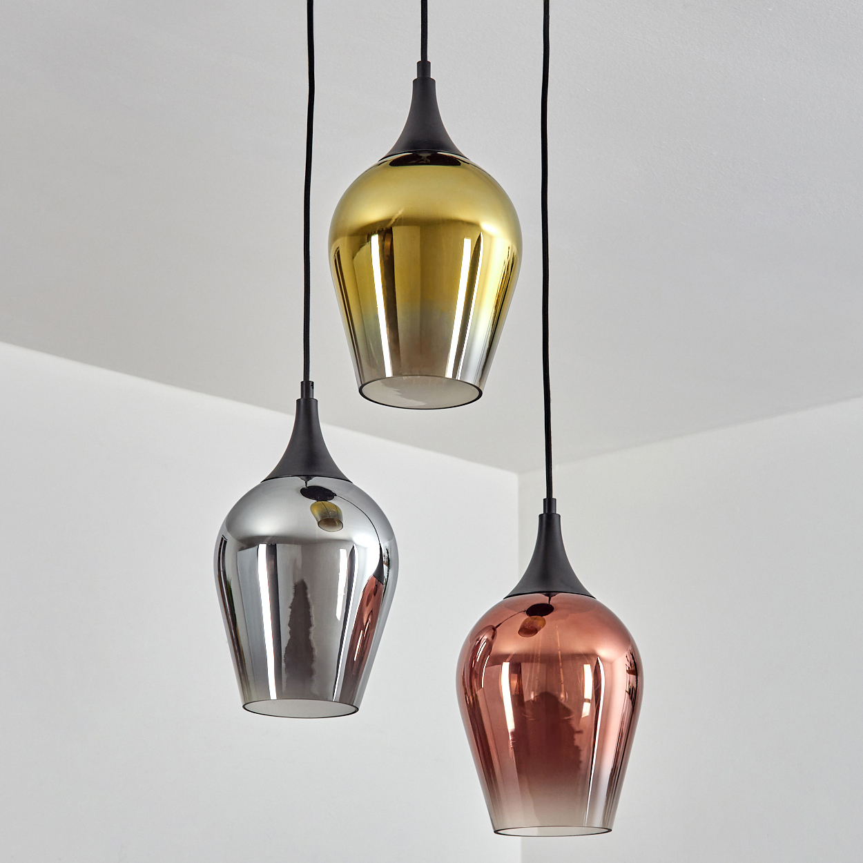 Drei Pendelleuchten mit metallischen Schirmen, Gold, Silber und Kupfer, hängen an schwarzen Kabeln von einer weißen Decke.