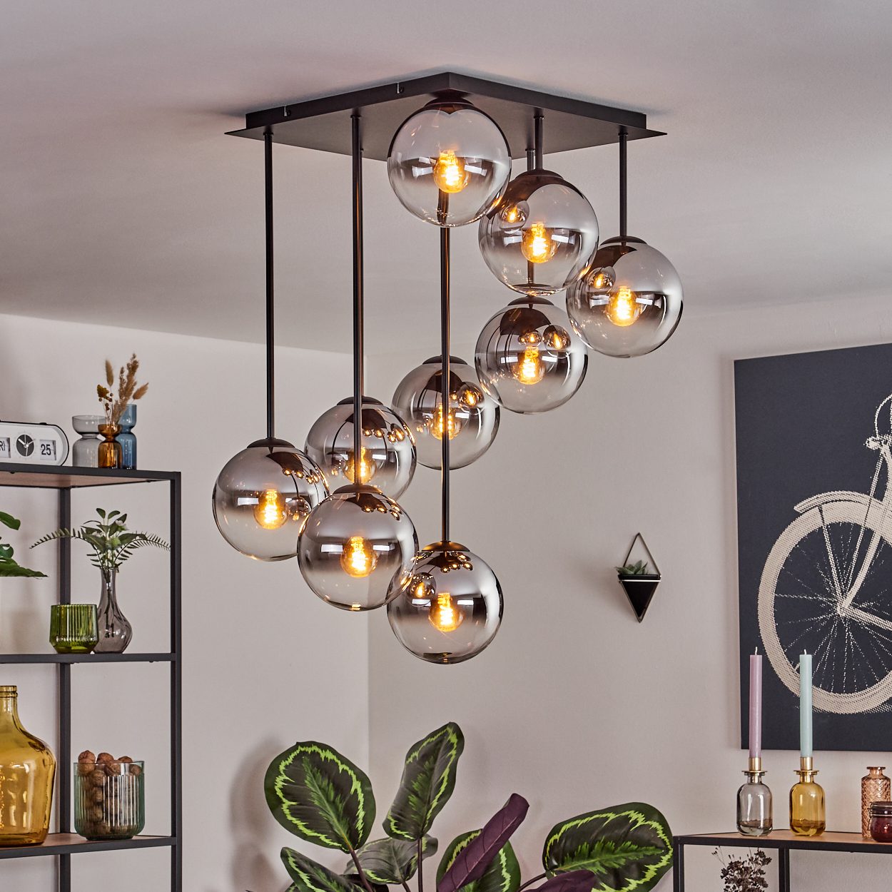 Eine moderne Deckenleuchte mit mehreren kugelförmigen Glaslichtern hängt von der Decke. Dekorative Gegenstände stehen auf Regalen und ein Wandbild.