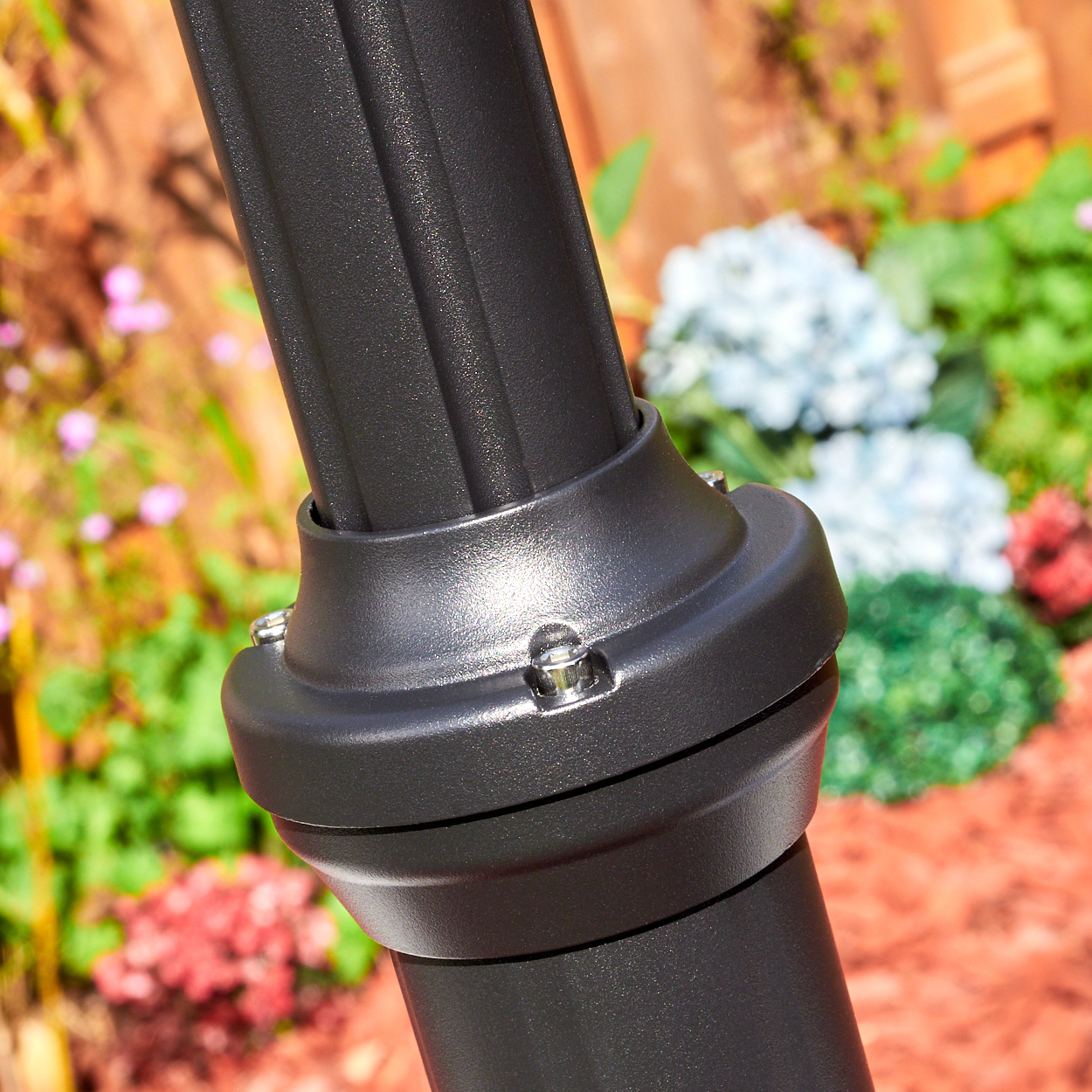 Nahaufnahme einer schwarzen Metallstange mit dekorativen Rippen. Eine abgerundete Basis mit Schrauben verbindet sich mit der Stange. Blumen und ein Zaun im verschwommenen Hintergrund.