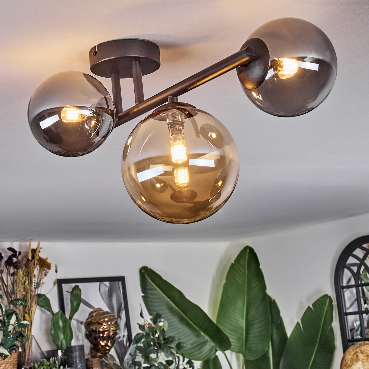 Eine moderne Deckenleuchte mit drei Glaskugeln. Das Licht ist an und wirft einen warmen Schein. Pflanzen und Dekor sind im Hintergrund.