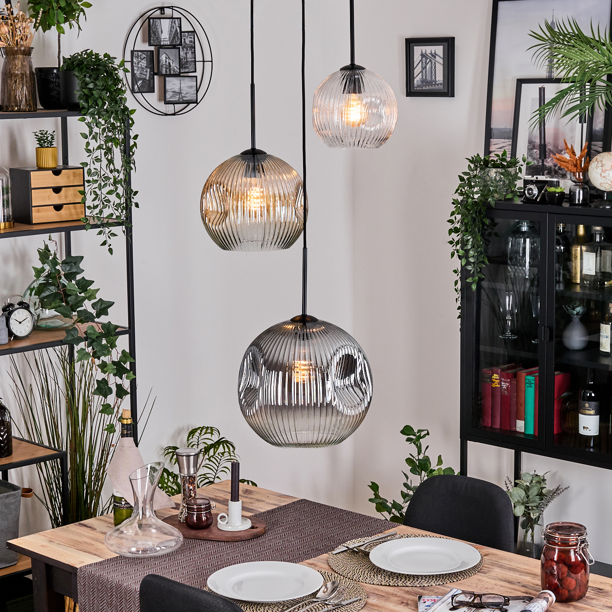 Esszimmer mit Holztisch, drei Hängelampen, Pflanzen, schwarzem Schrank und gerahmten Bildern. Teller sind gedeckt.