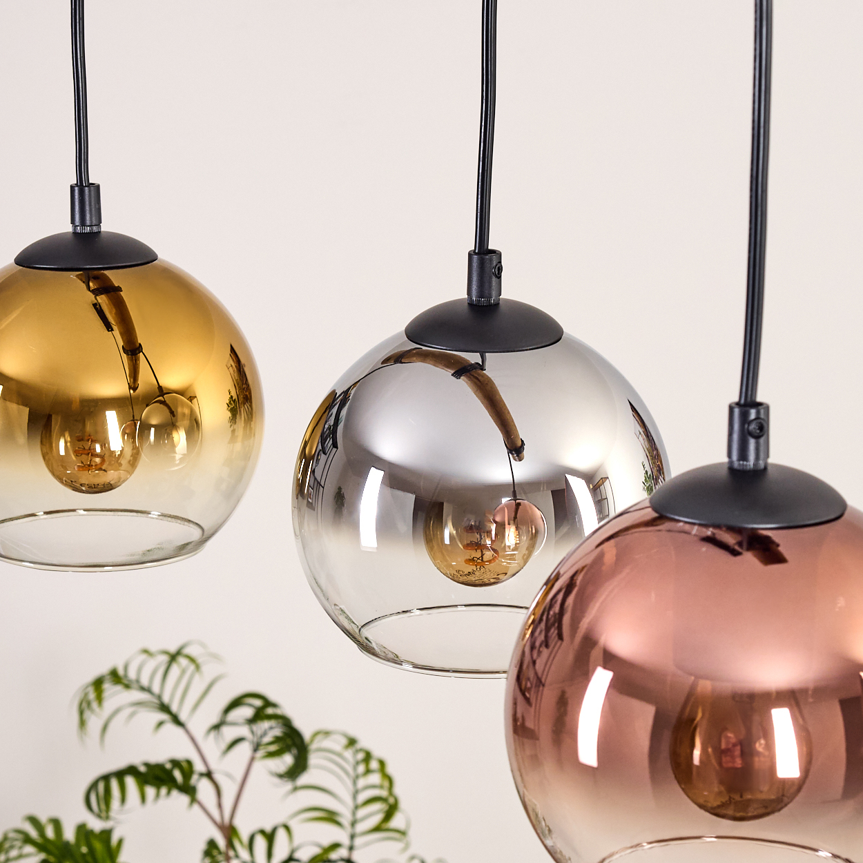 Drei kugelförmige Pendelleuchten in Gold, Silber und Kupfer. Jede hat ein schwarzes Kabel und eine sichtbare Glühbirne. Grüne Pflanze im Hintergrund.