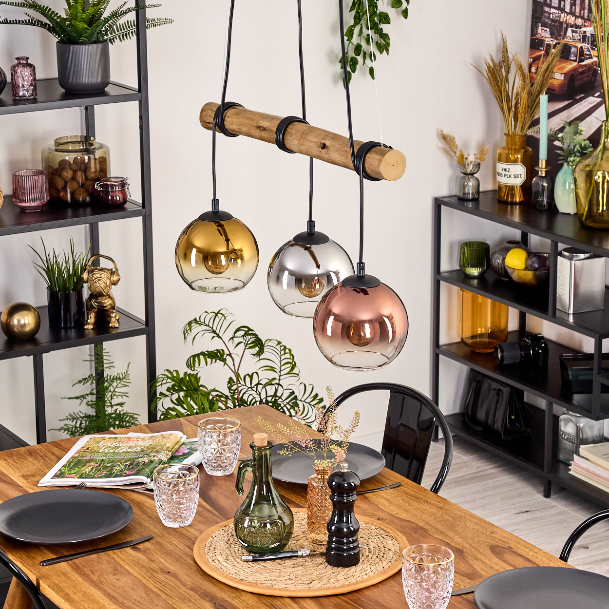 Esszimmer mit Holztisch, drei Hängelampen, schwarzen Regalen mit Pflanzen und Dekorationen und einer Zeitschrift.
