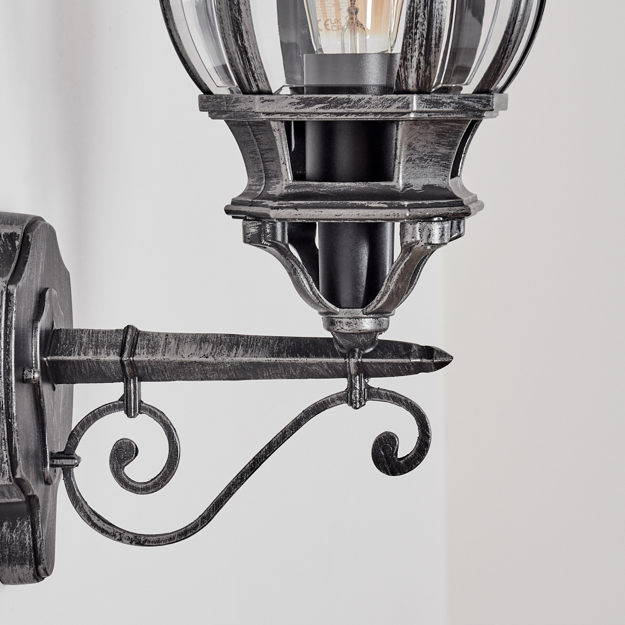 Eine dekorative Wandleuchte. Die Lampe hat einen klaren Glasschirm mit einem silbernen Metallrahmen. Die Halterung ist verziert, dunkles Metall.