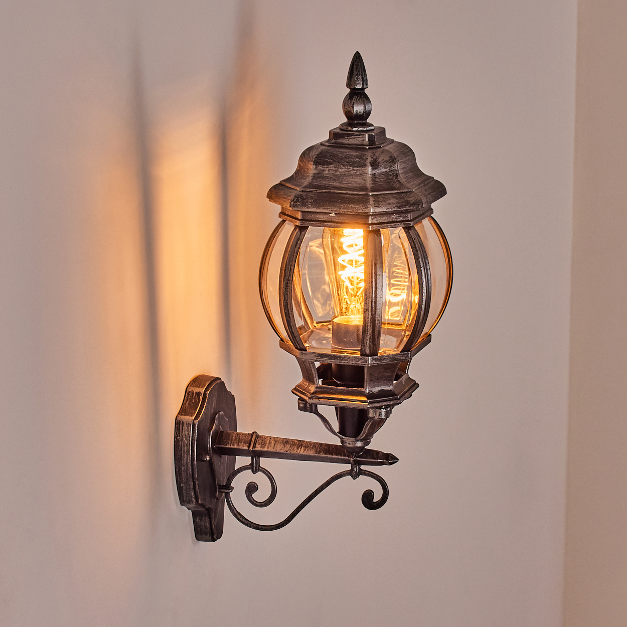 Eine verzierte Wandlampe mit leuchtender Glühbirne. Sie hat eine Bronzefinish und ist an einer hellen Wand befestigt, die einen Schatten wirft.