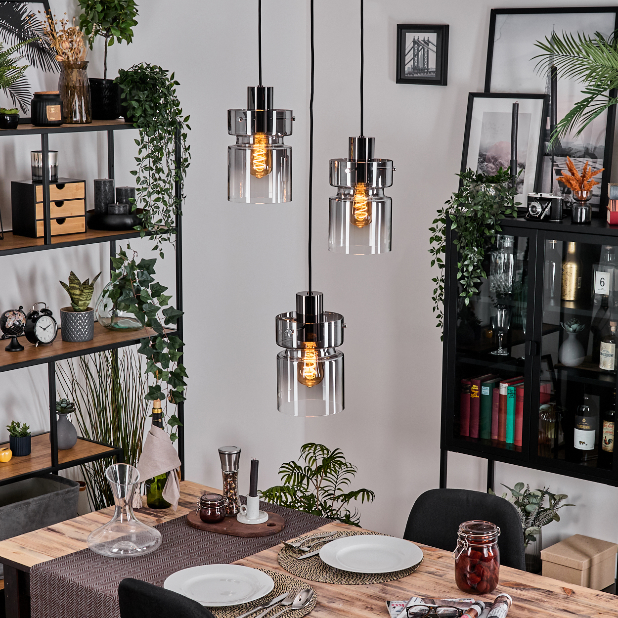 Esszimmer mit Holztisch, drei Hängelampen, Pflanzen und Regalen. Teller, Besteck und Dekorationen stehen auf dem Tisch.