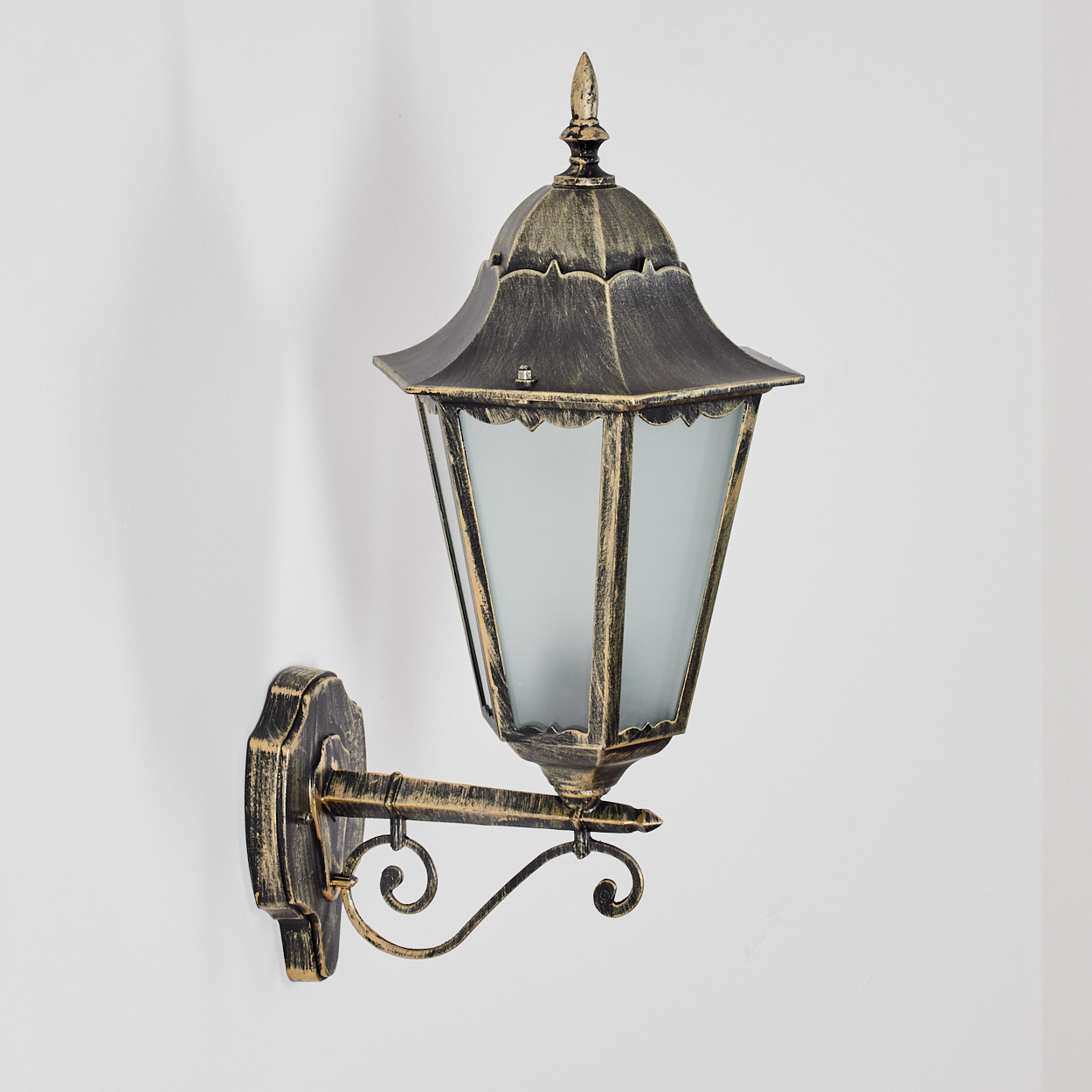 Eine verzierte, bronzefarbene Wandlampe mit Glaspaneelen ist an einer weißen Wand befestigt. Die Lampe hat ein dekoratives Design und eine spitze Spitze.
