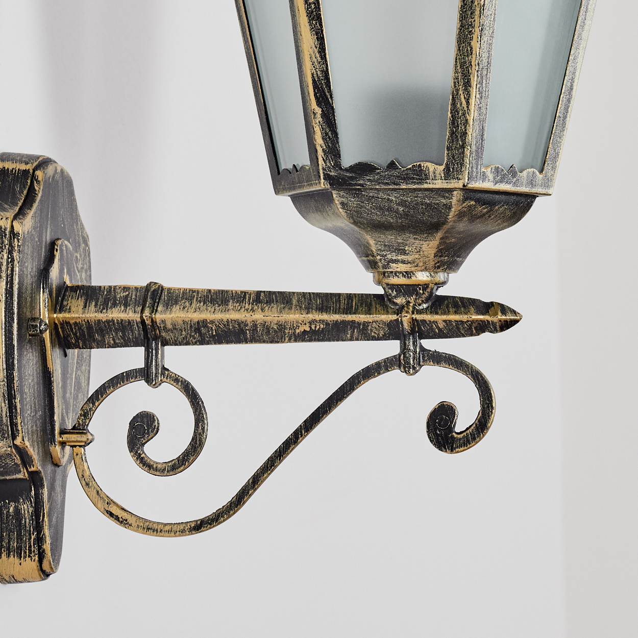 Eine dekorative Wandlampe mit schwarzer und goldener Oberfläche. Die Lampe hat einen Glasschirm und Ziergitter aus Metall, an einer weißen Wand montiert.