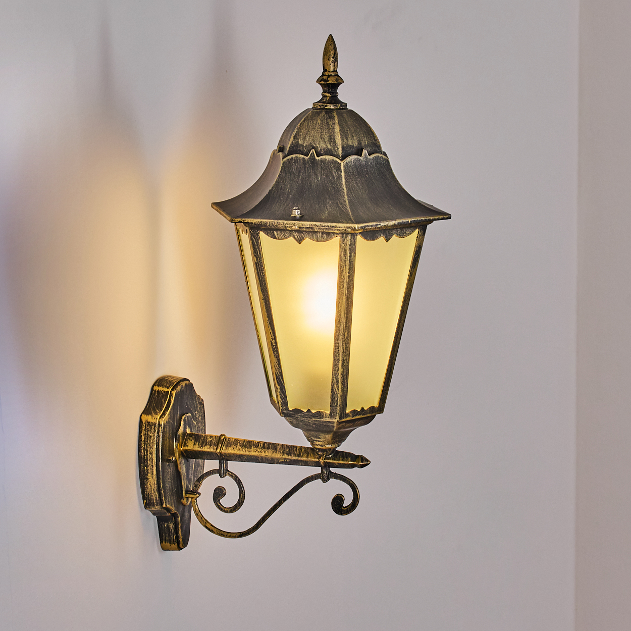 Eine verzierte Wandlampe mit goldenem Finish ist an einer weißen Wand befestigt. Die Lampe hat ein gelbes Licht und ist eingeschaltet.
