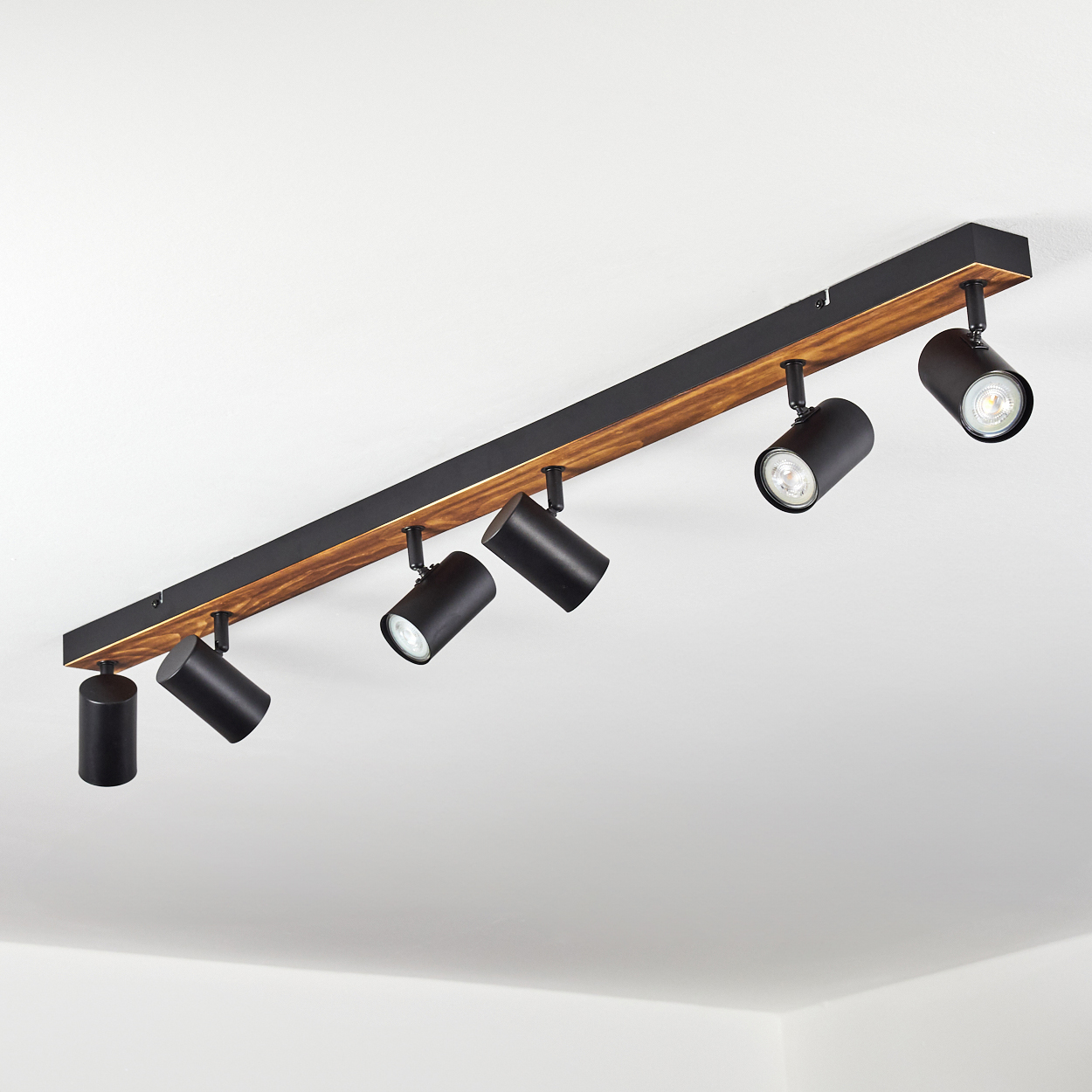 Eine Deckenleuchte mit sechs schwarzen Strahlern, die an einem Holz- und schwarzen Metallbalken befestigt sind, vor weißer Decke.