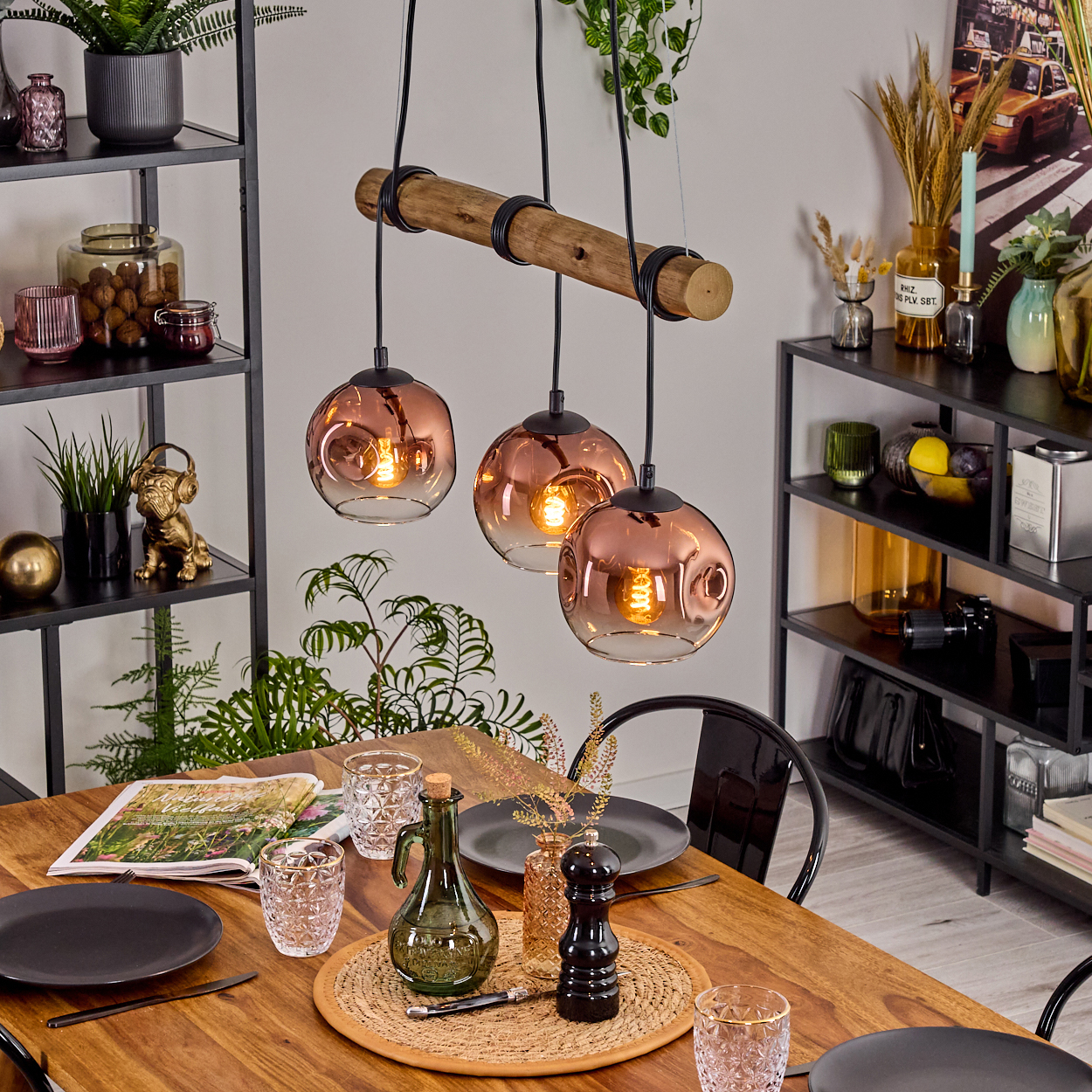 Esszimmer mit Holztisch, drei Hängelampen, schwarzen Regalen mit Dekor und einer Tischdekoration mit Tellern und Gläsern.