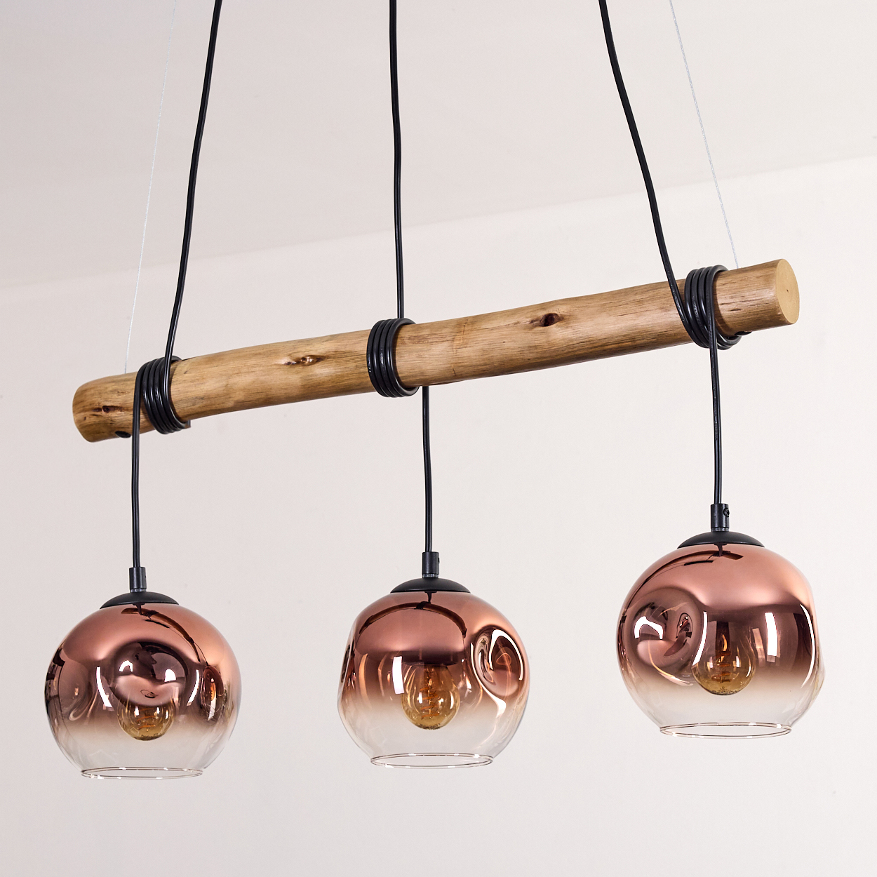 Drei kupferfarbene Glas-Pendelleuchten hängen an einem Holzbalken. Jede hat eine Glühbirne. Schwarze Schnüre und Ringe sind sichtbar.