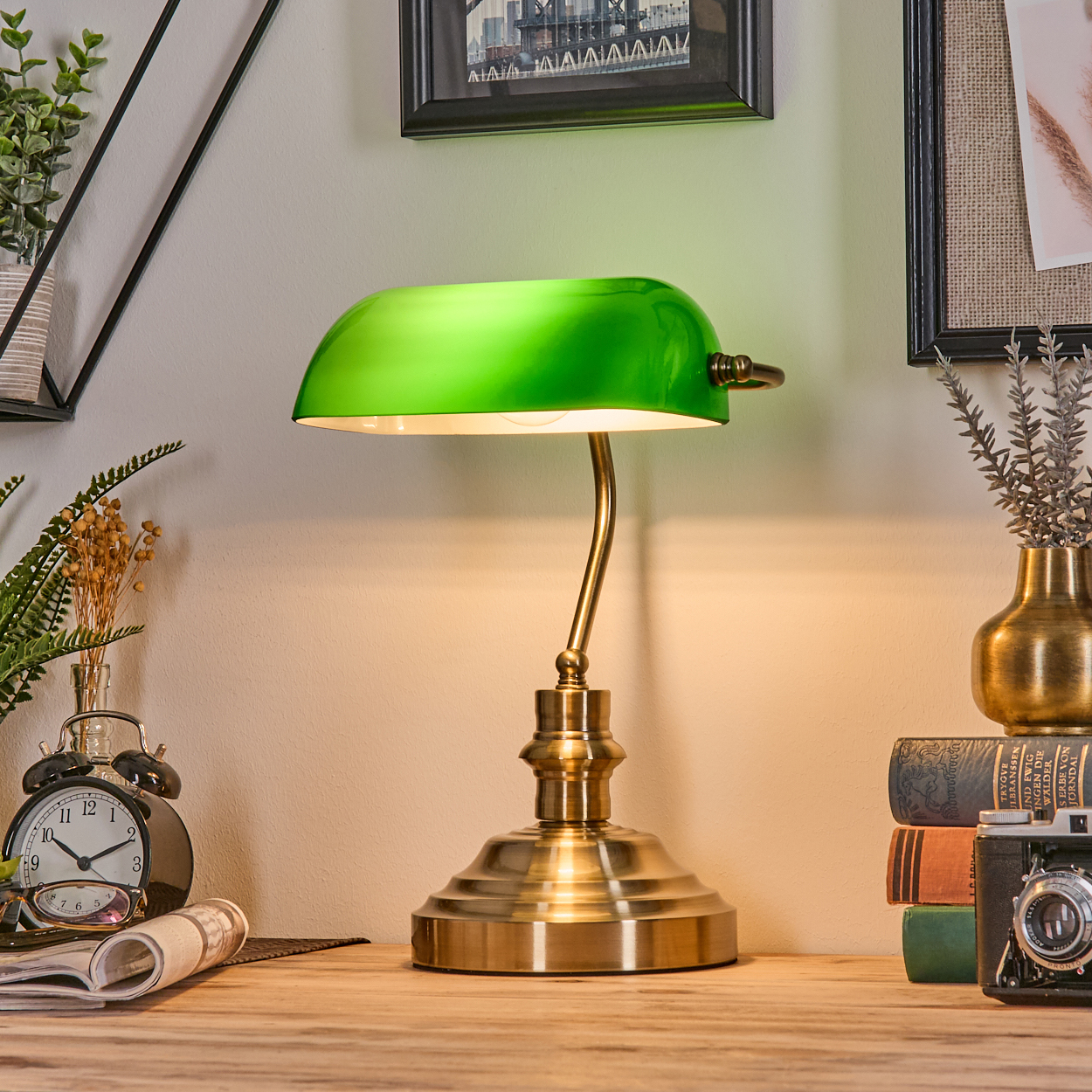 Eine grüne Bankerlampe steht auf einem Holztisch mit Büchern, einer Kamera und einer Uhr. Im Hintergrund sind Bilder und Pflanzen.