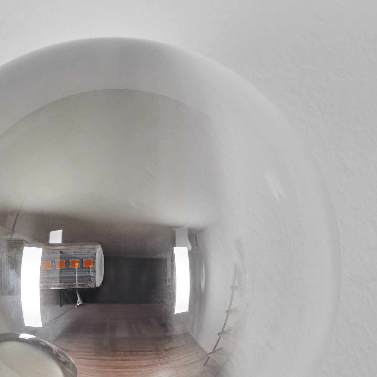 Ein kugelförmiges Glasobjekt spiegelt einen Raum mit Tisch, Lichtern und einer Leiter an einer weißen Wand wider.