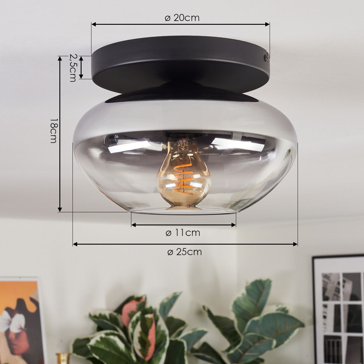 Eine moderne Deckenlampe mit schwarzem Sockel und Glasschirm, mit Maßangaben. Pflanzen und ein gerahmtes Bild im Hintergrund.