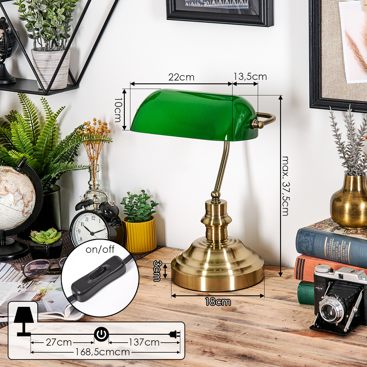 Eine grüne Schreibtischlampe aus Messing steht auf einem Holztisch mit einem schwarzen Ein-/Ausschalter. Weitere Objekte sind Bücher, eine Kamera und Pflanzen.