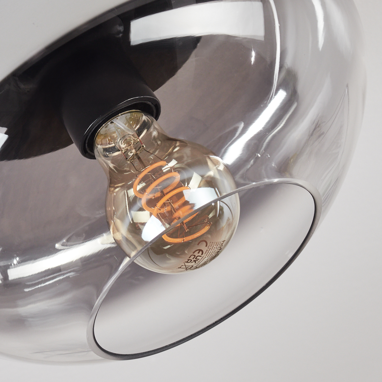 Nahaufnahme einer Leuchte, die eine Glühbirne mit gewendeltem Glühfaden in einem transparenten Glasschirm zeigt. Die Halterung ist schwarz und silber.