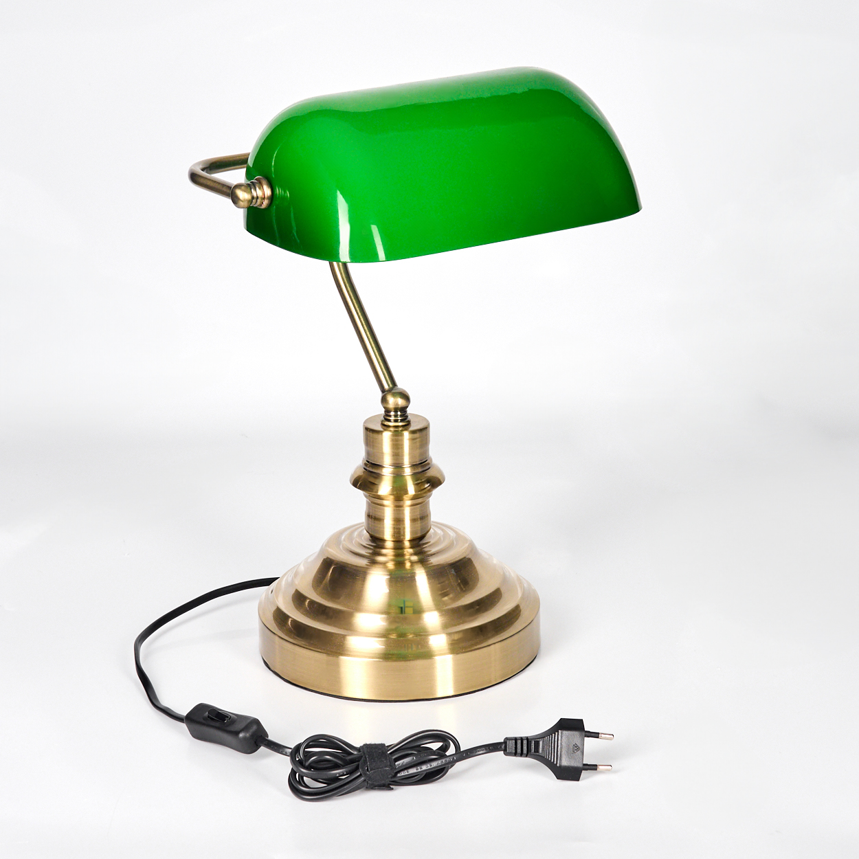 Eine grüne Schreibtischlampe aus Messing steht auf einer weißen Oberfläche. Das Kabel der Lampe ist schwarz und hat einen Stecker.