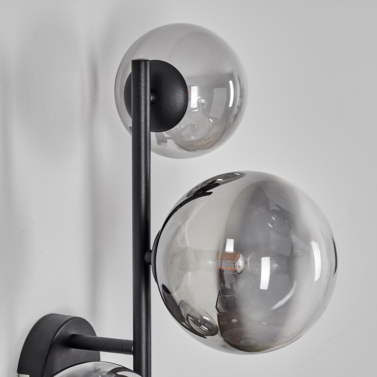 Eine moderne Wandlampe mit drei grauen Glaskugeln. Schwarzer Metallrahmen und Halterung, an einer weißen Wand befestigt. Die Kugeln reflektieren das Licht.