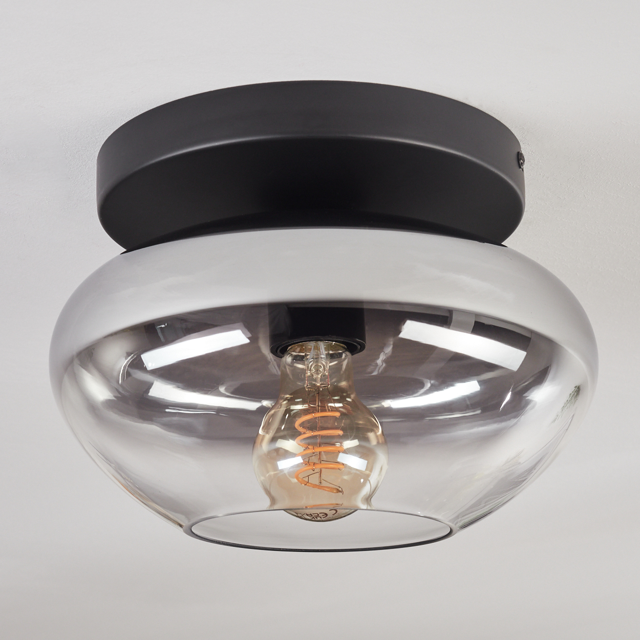 Eine moderne Deckenleuchte. Sie hat einen schwarzen Sockel, einen silbernen Rand und einen transparenten, schalenförmigen Glasschirm.