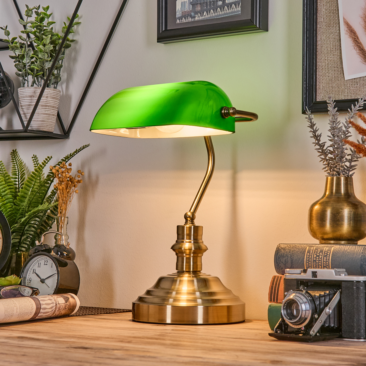 Eine grüne Banker-Lampe steht auf einem Holztisch mit einer Kamera, Büchern und einer Vase mit trockenen Blumen. Pflanzen und eine Uhr sind links.