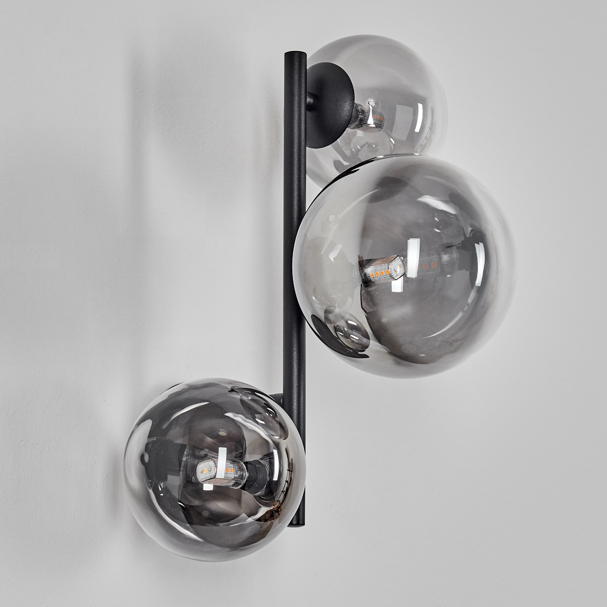 Eine moderne Wandleuchte mit drei kugelförmigen, rauchfarbenen Glasschirmen. Die Halterung ist schwarz und an einer weißen Wand montiert.