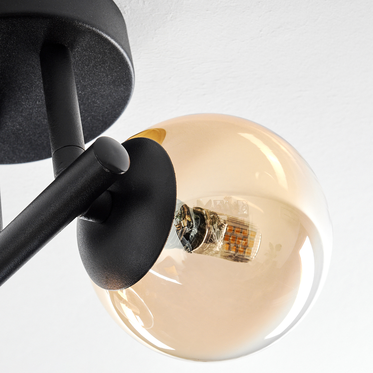 Nahaufnahme einer schwarzen Deckenlampe mit rundem, bernsteinfarbenem Glasschirm. Die Glühbirne ist sichtbar. Weißer Hintergrund.