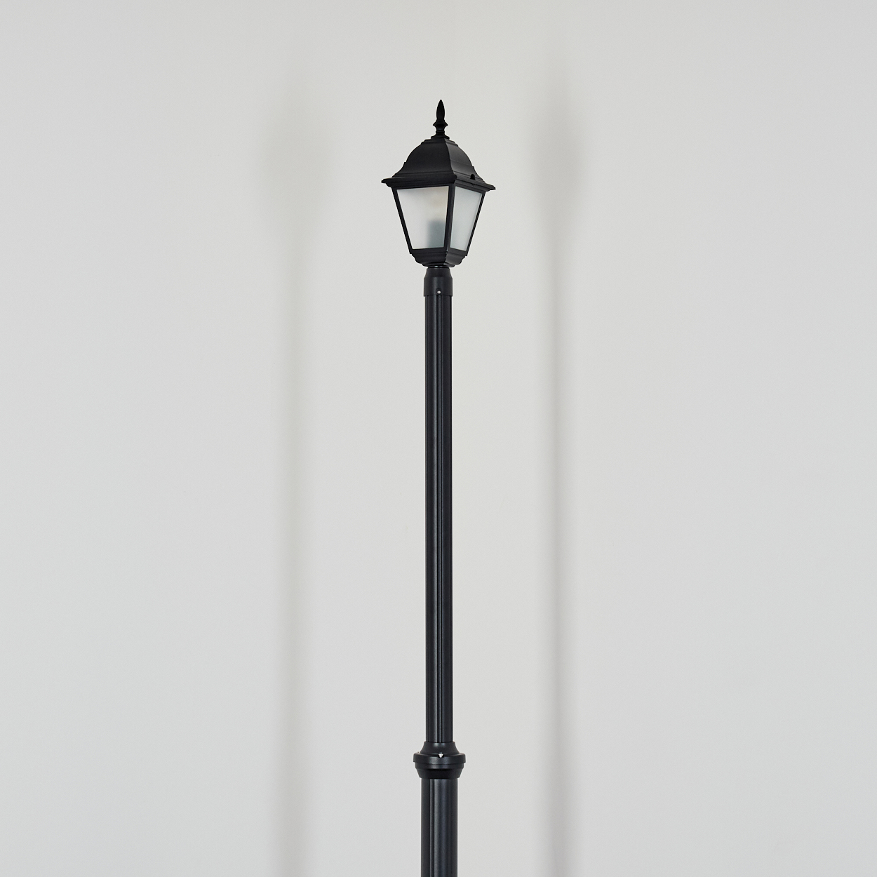 Eine schwarze Straßenlaterne steht vor einer weißen Wand. Die Laterne hat eine dekorative Spitze und ein Licht. Ein Schatten wird geworfen.