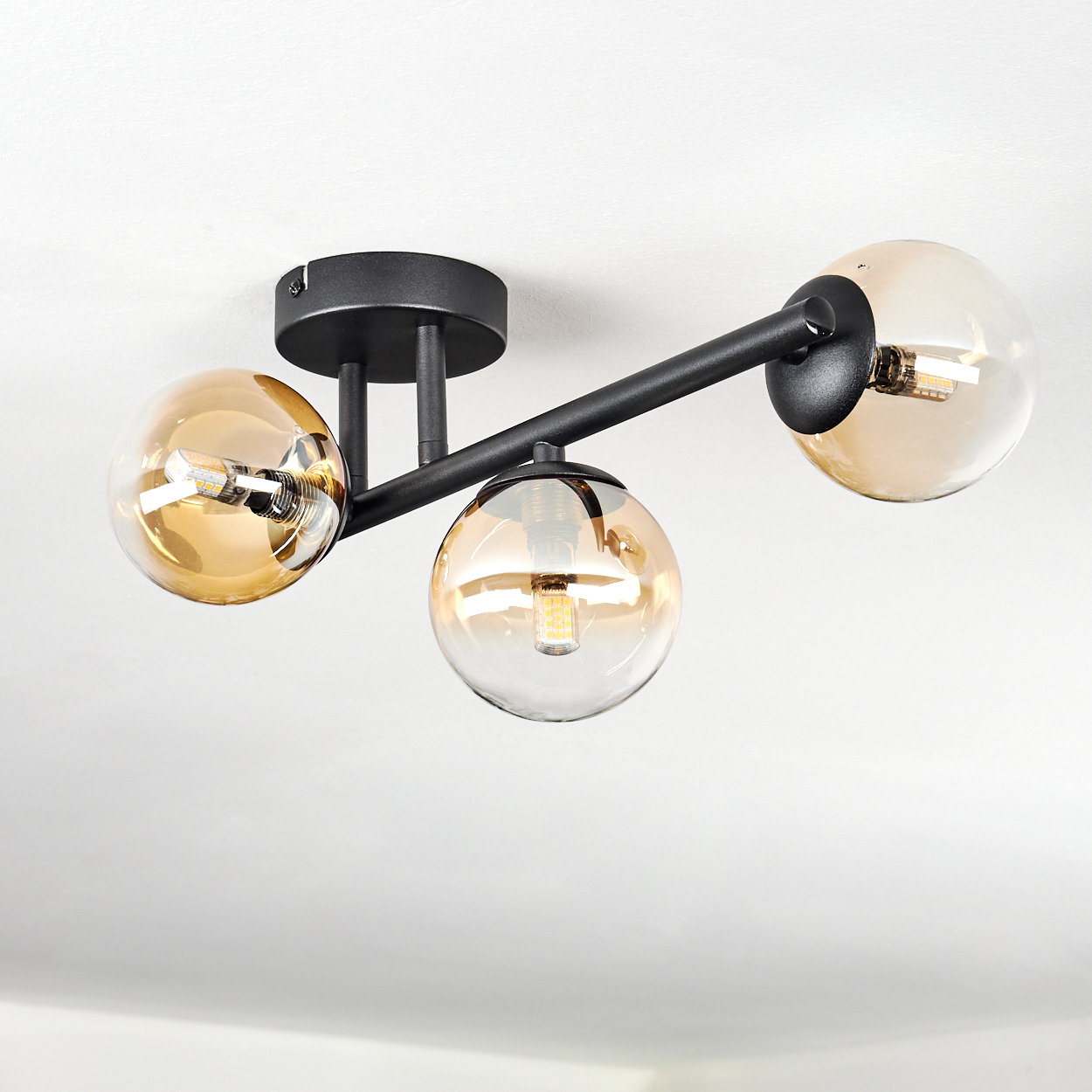 Deckenlampe mit drei bernsteinfarbenen Glaskugeln und schwarzen Metallarmen, jede mit einer Glühbirne. Sie ist vor einem weißen Hintergrund.