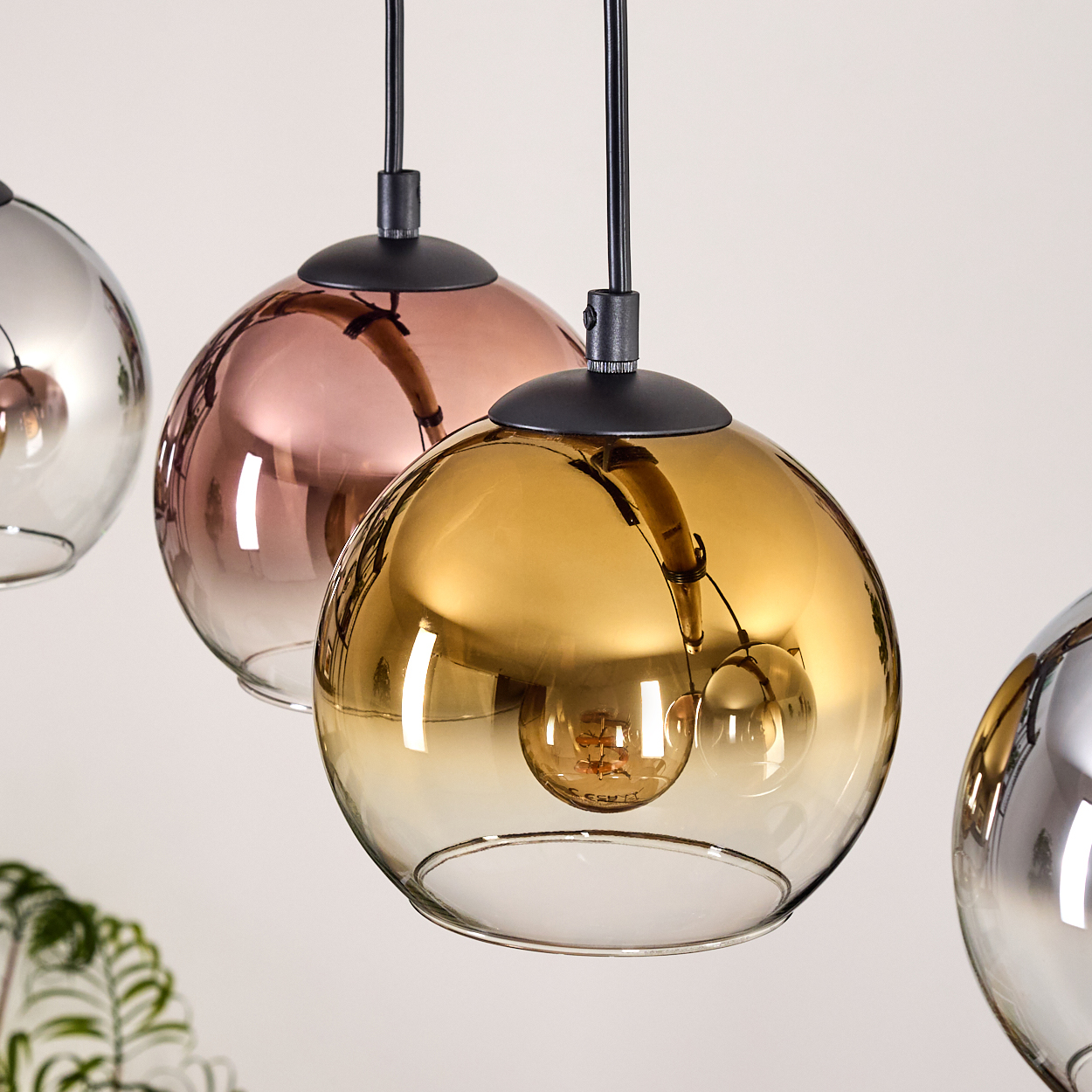 Vier kugelförmige Pendelleuchten mit Farbverläufen: Silber, Kupfer, Gold. Schwarze Kabel und Armaturen. Schlichter weißer Hintergrund und Pflanze.