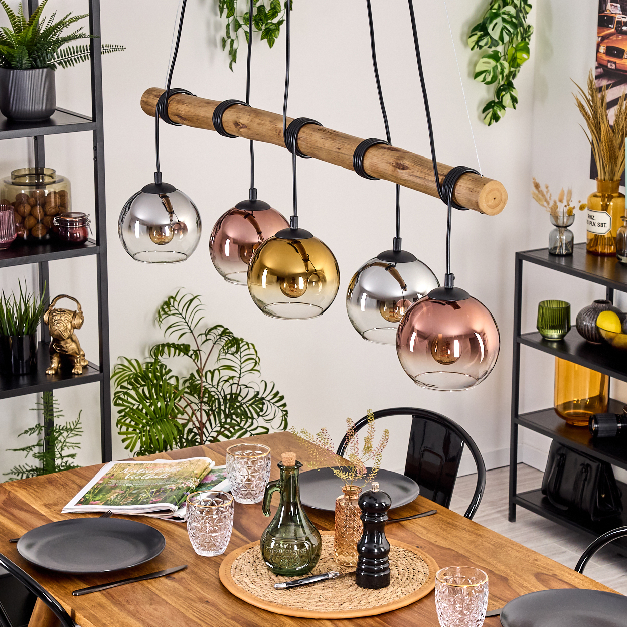Ein Esszimmer mit Holztisch, Hängelampen und Tischdeko. Schwarze Regale und Pflanzen sind im Hintergrund zu sehen.