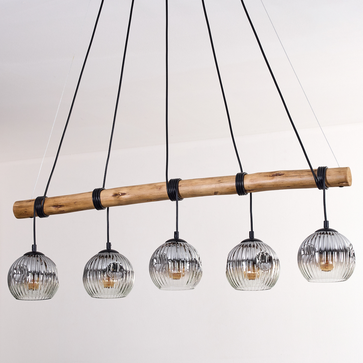 Eine moderne Hängeleuchte mit fünf Glaskugelleuchten hängt von einem Holzbalken. Schwarze Kabel und Ringe verbinden die Leuchten mit dem Balken.