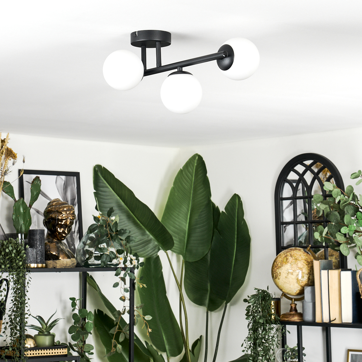 Ein modernes Zimmer mit einer schwarz-weißen Deckenlampe, Pflanzen und einem schwarzen Regal mit dekorativen Gegenständen. Der Raum ist gut beleuchtet.