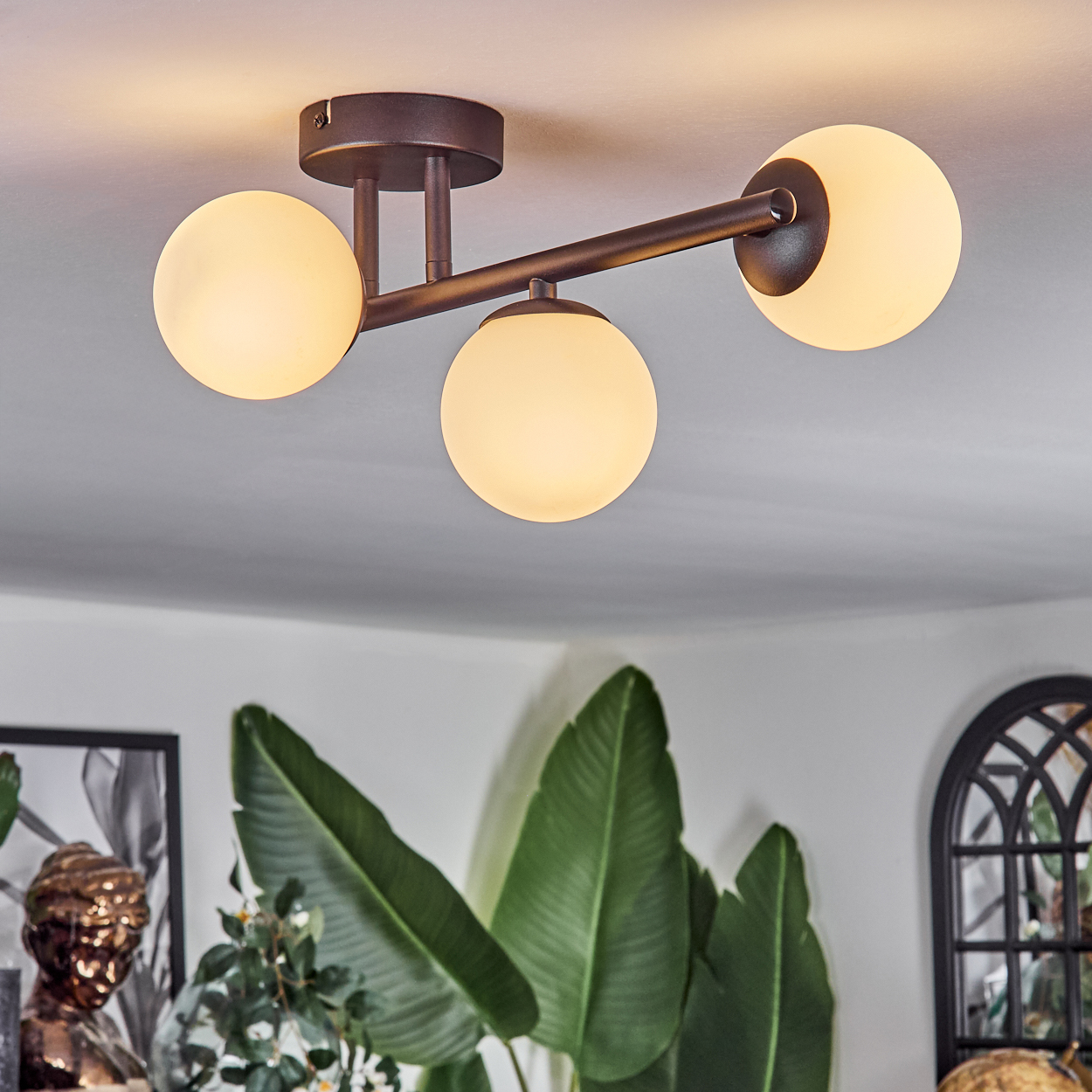 Eine moderne Deckenleuchte mit drei kugelförmigen Lampen. Darunter sind große grüne Blätter und eine Büstenskulptur zu sehen.