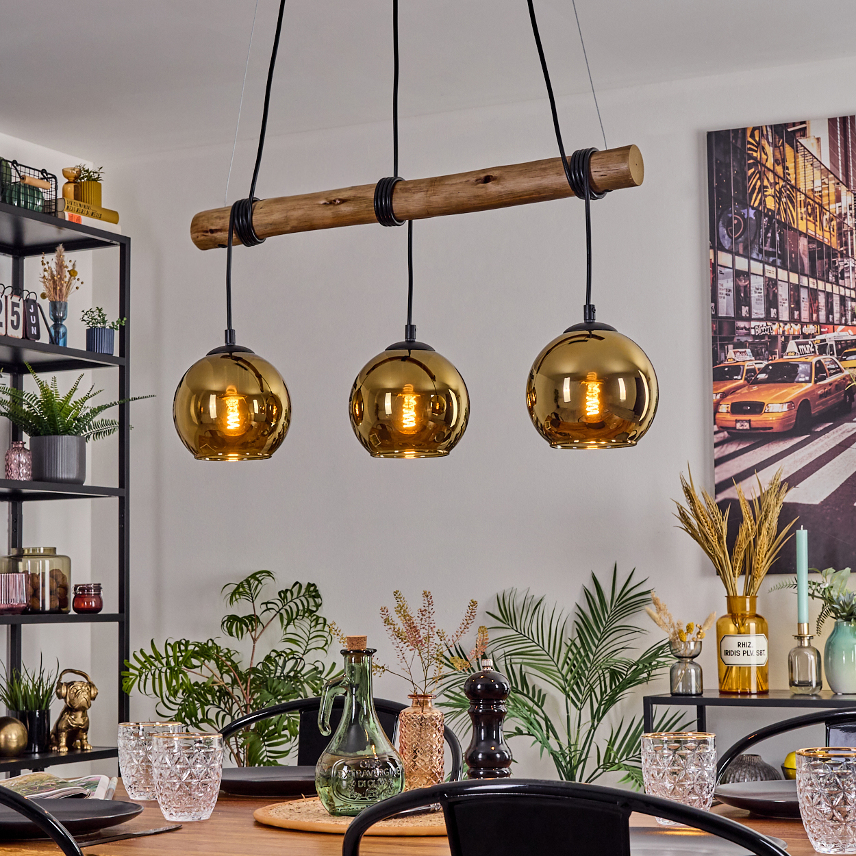 Drei goldene Pendelleuchten über einem Esstisch. Im Hintergrund ein Gemälde einer Stadtszene, Pflanzen und Regale.